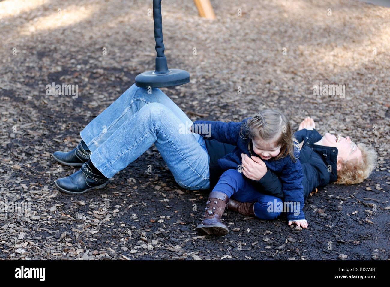 A middle aged woman, laughing out loud, whilst lying on the ground after falling off play apparatus she had been using with her Grandchild Stock Photo