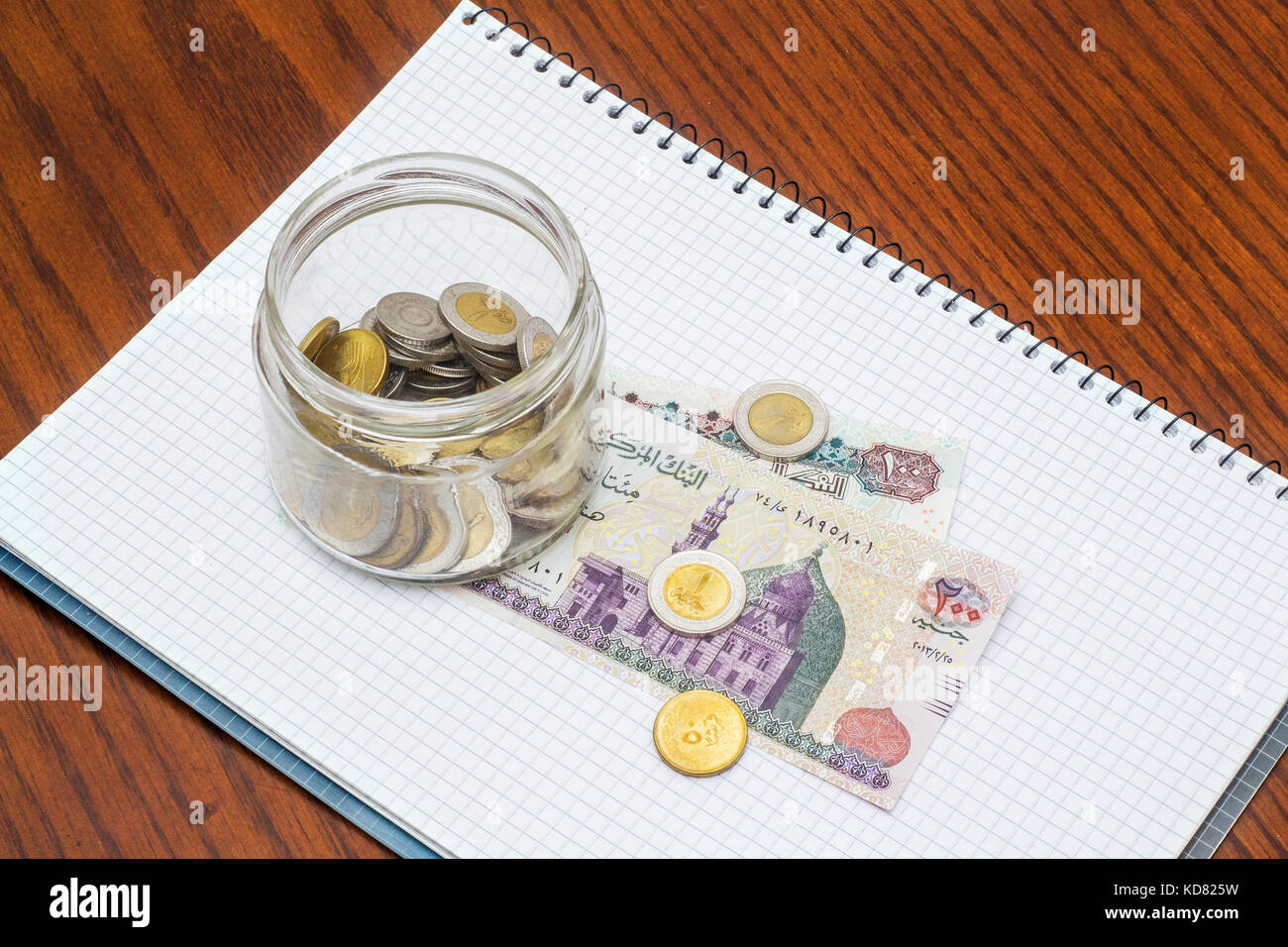 Glass Full of Coins on Banknotes with Notebook, on Wooden Desk Stock Photo