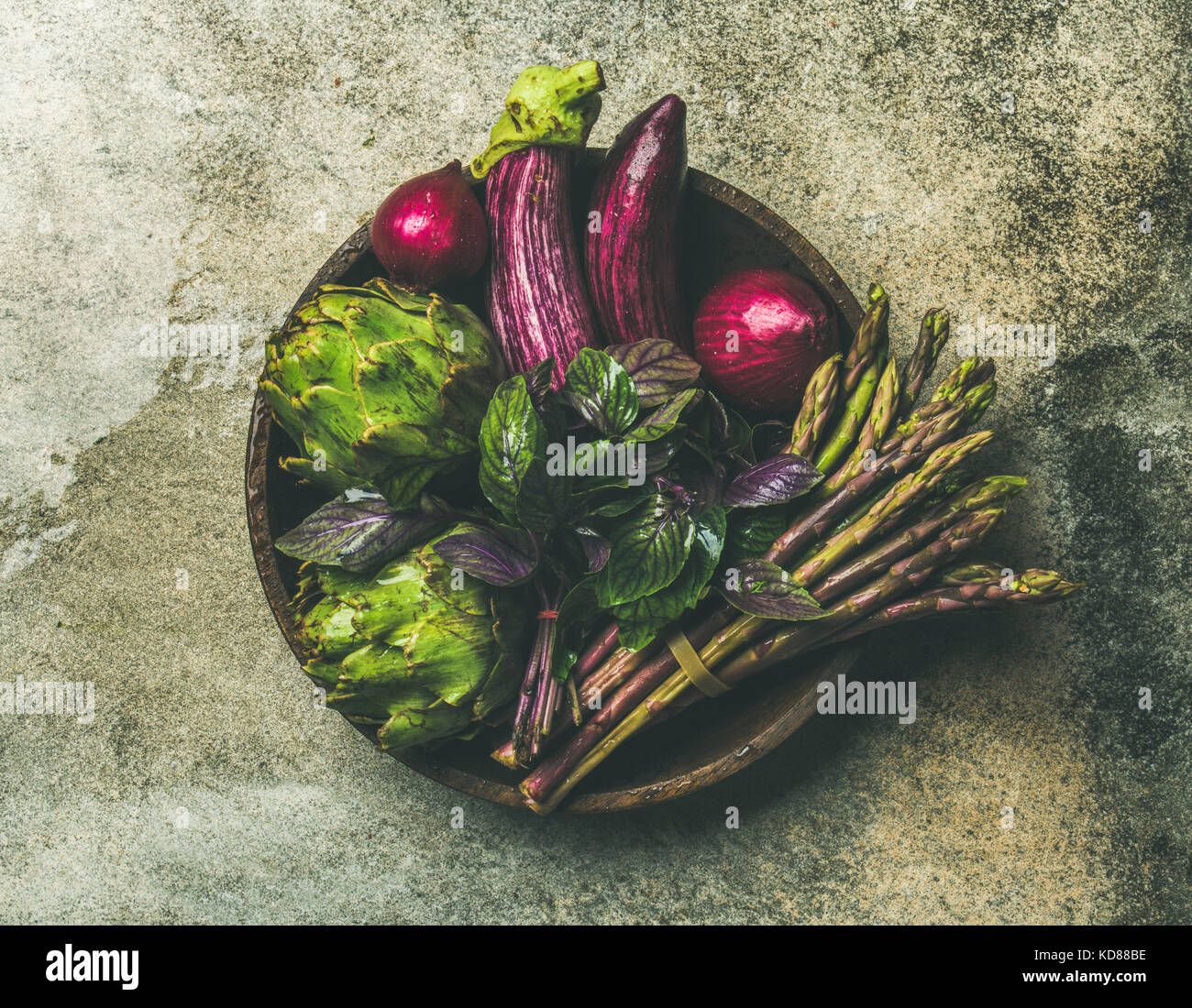 Flat-lay of green and purple vegetables on plate over grey background, top view. Local produce for healthy cooking. Eggplans, beans, kale, asparagus, Stock Photo