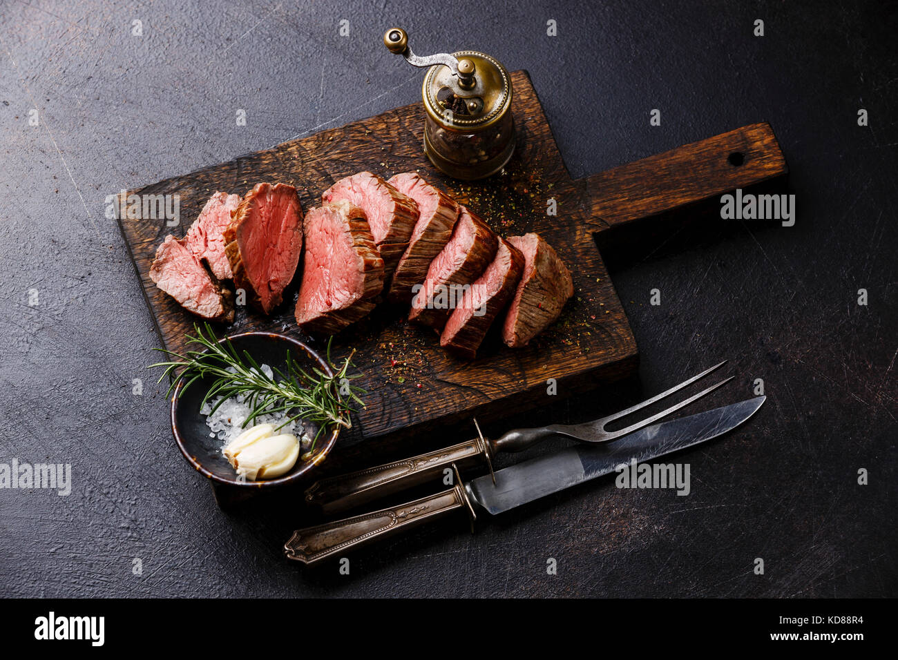 Sliced tenderloin Steak Roast beef with knife and fork carving set on wooden cutting board on dark background Stock Photo