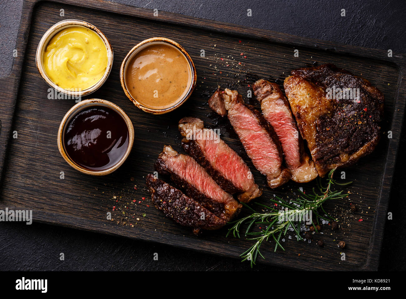 Sliced grilled Rib eye steak and three different sauces: Pepper sauce, Mustard and Barbecue on dark background close up Stock Photo