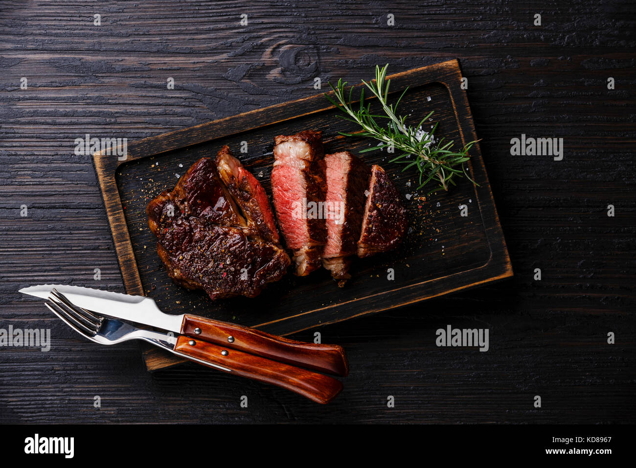 Sliced grilled meat barbecue steak Rib eye with knife and fork on burned black wooden background Stock Photo