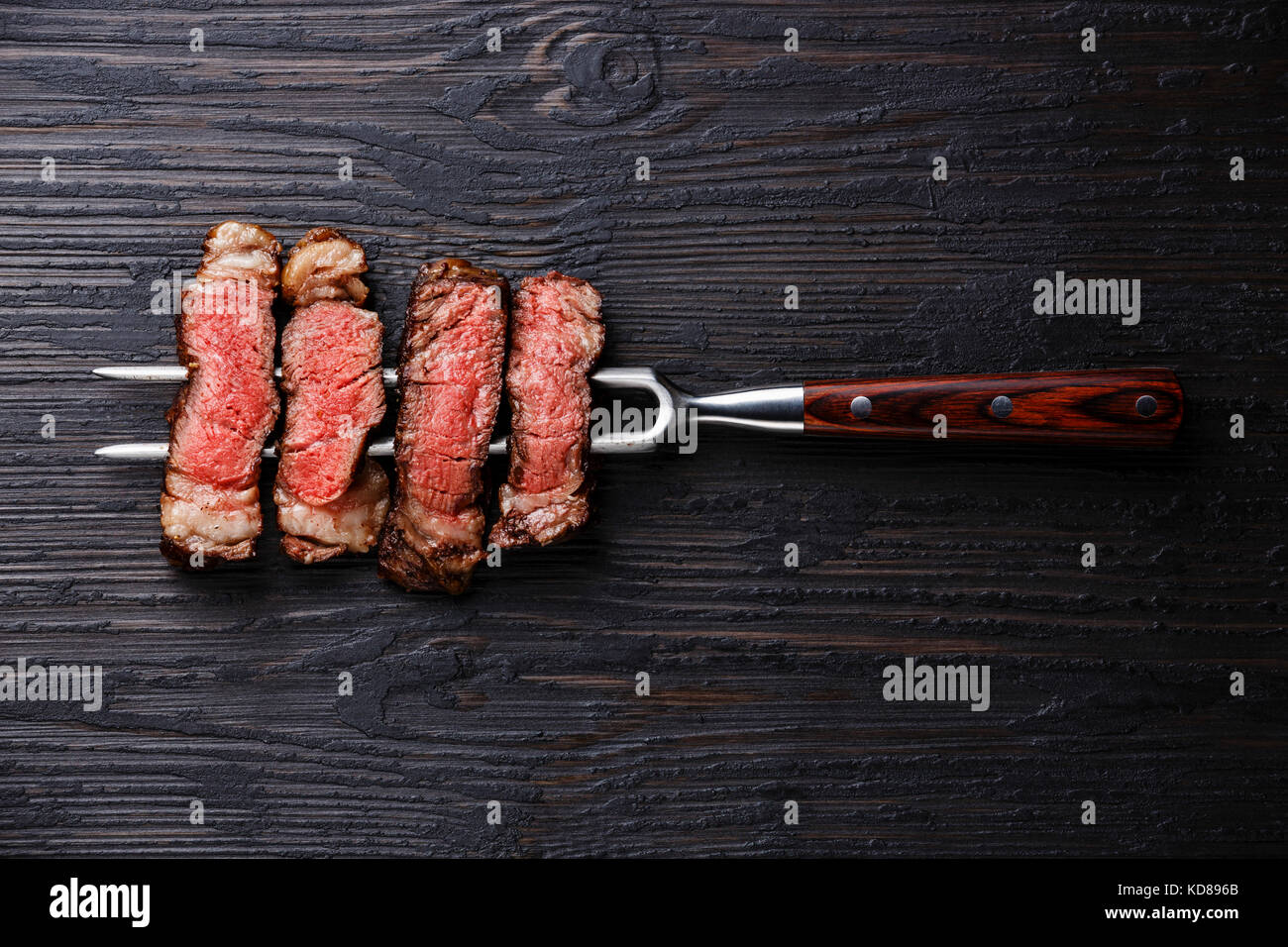 Slices of grilled meat barbecue steak Rib eye on meat fork on burned black wooden background Stock Photo
