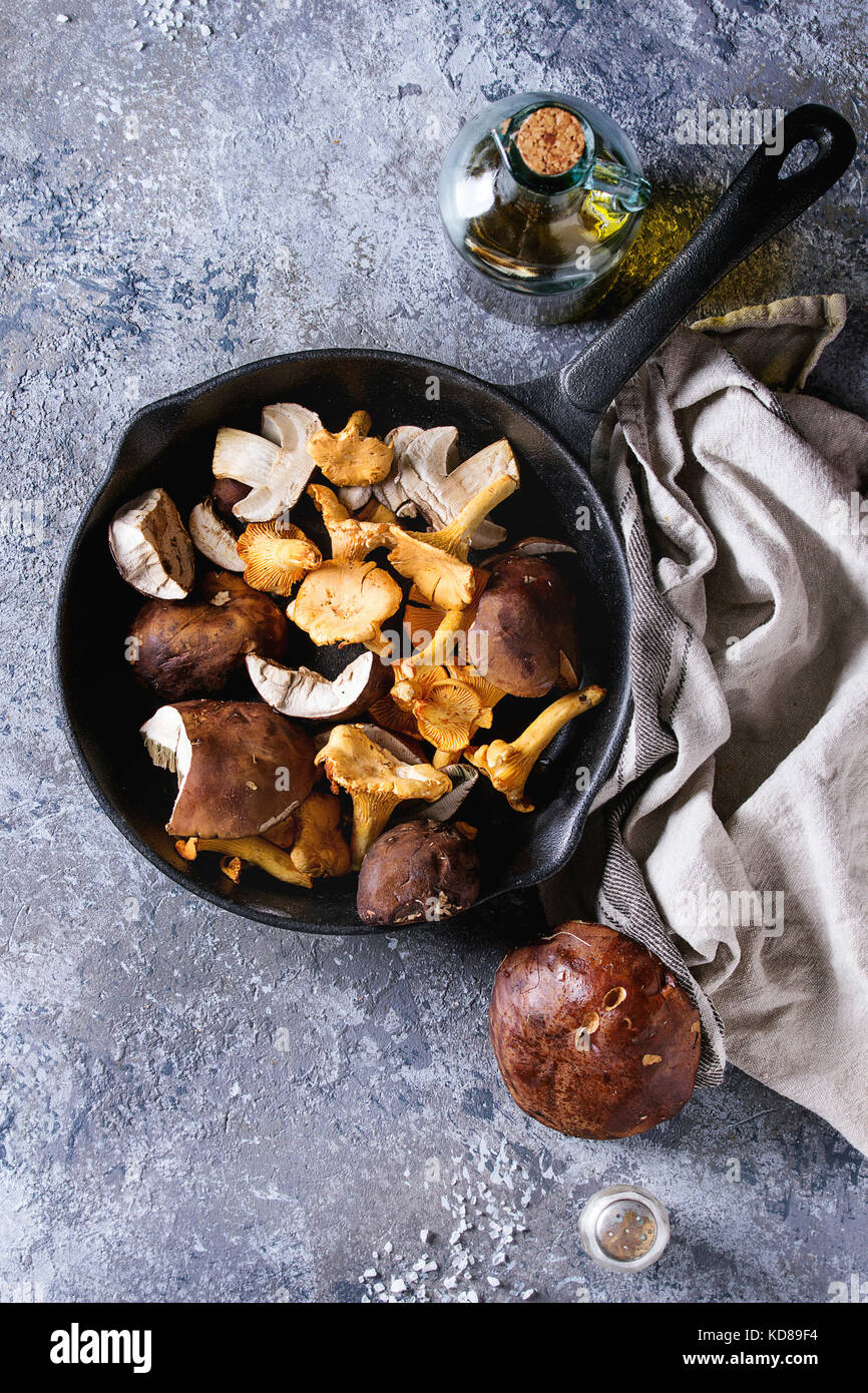 Heap of fresh forest porcini boletus and chanterelles mushrooms in iron cast pan with olive oil and salt, served won textile napkin with knife over gr Stock Photo