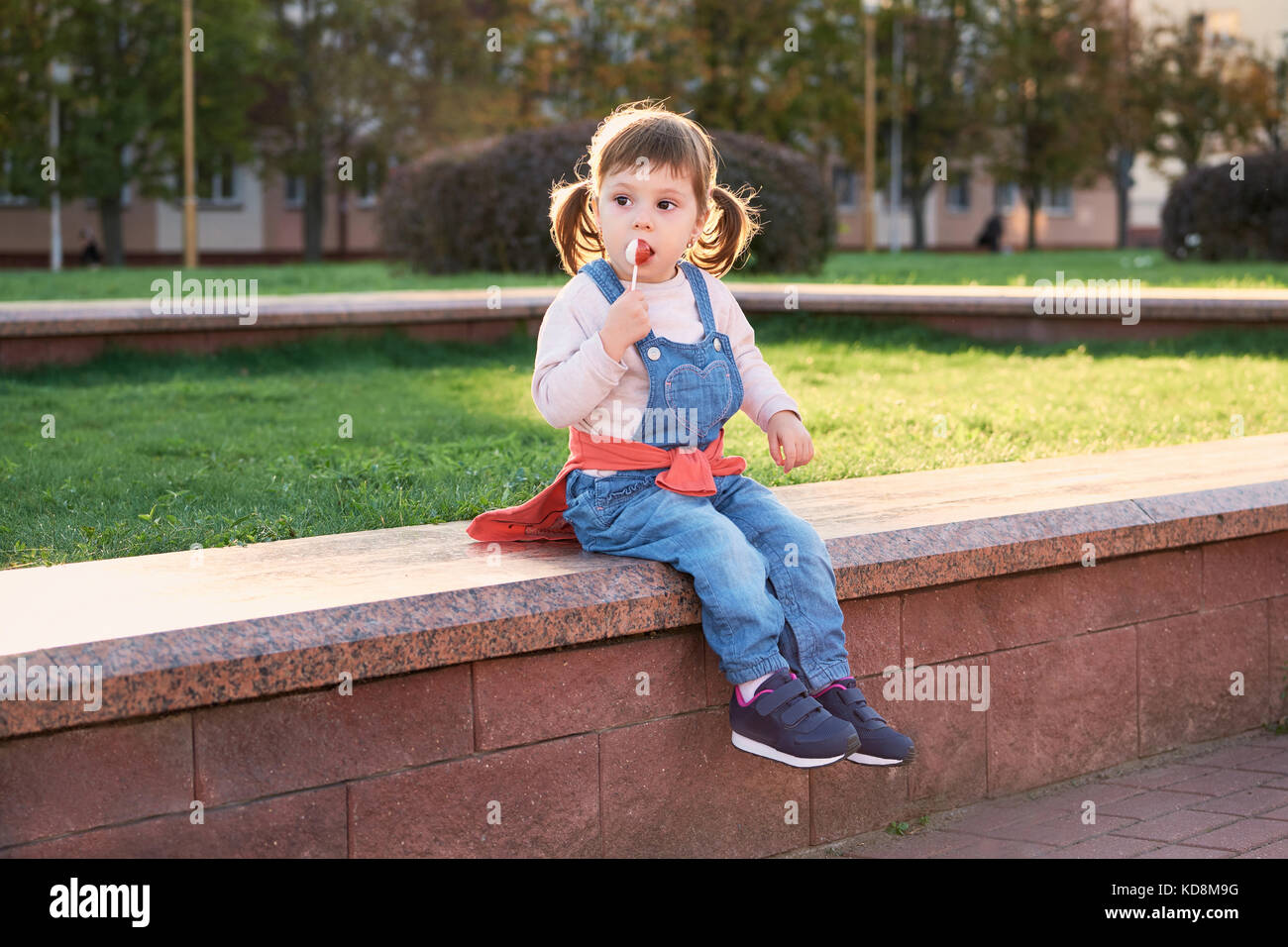 Cute beautiful child eats candy.little girl in denim jumpsuit Stock