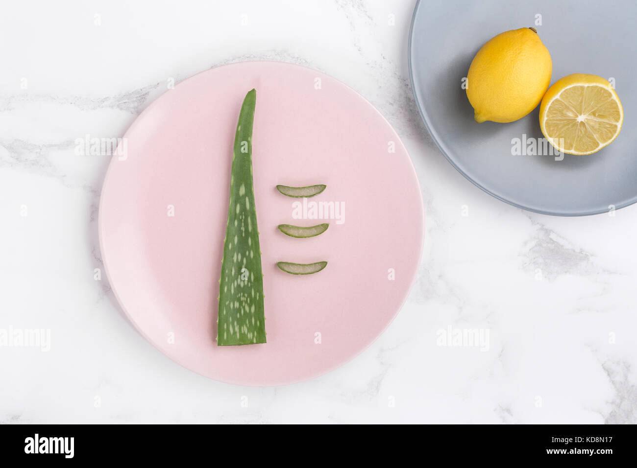 Styled lifestyle minimalist stock photo of an aloe vera plant leaf and cut open lemons on pink and blue plates on a marble background. Stock Photo