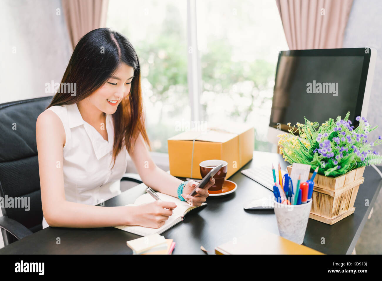 Young Asian small business owner work at home office, using mobile phone call, write purchase order on notebook. financial or entrepreneur concept Stock Photo