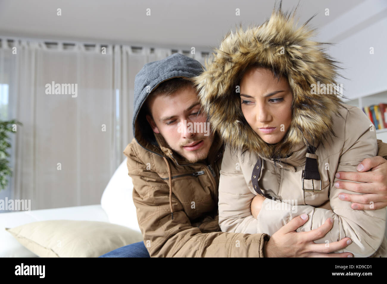 Cold home with an angry couple warmly clothed hugging sitting on a sofa in the living room Stock Photo