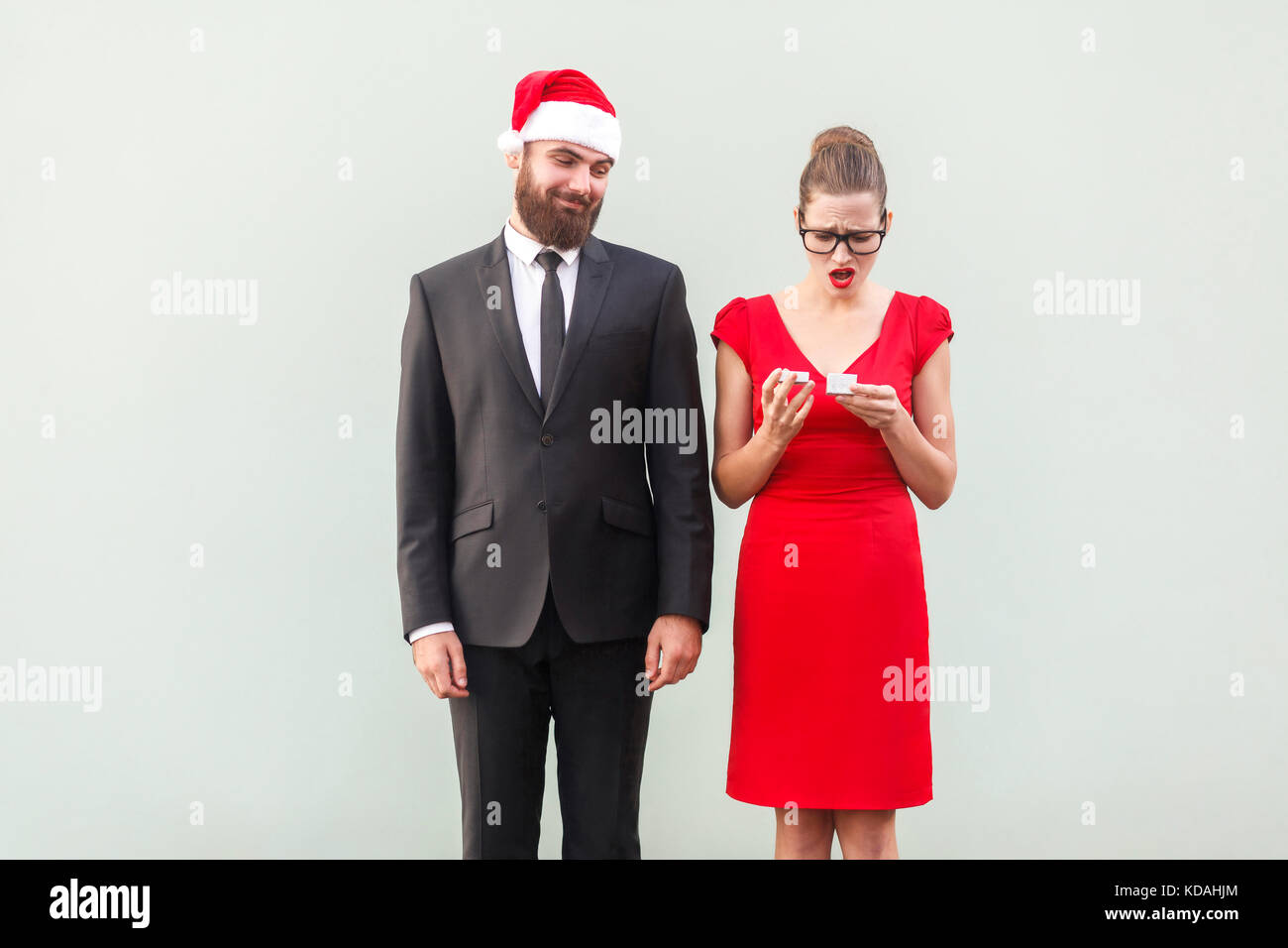 Proud man in christmas cap looking a gift. Frustrated woman holding gifts. Studio shot, gray wall Stock Photo