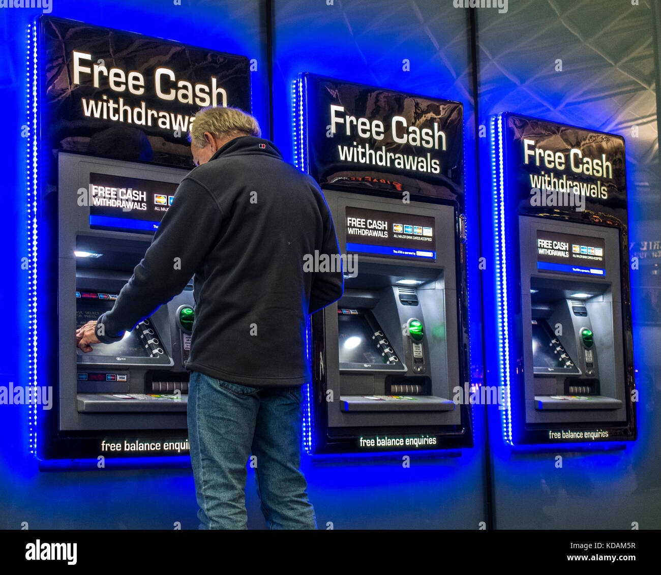 Man making cash machine / cashpoint / ATM withdrawal at a row of three machines, with glowing blue lighting. Kings Cross Station, London, England, UK. Stock Photo