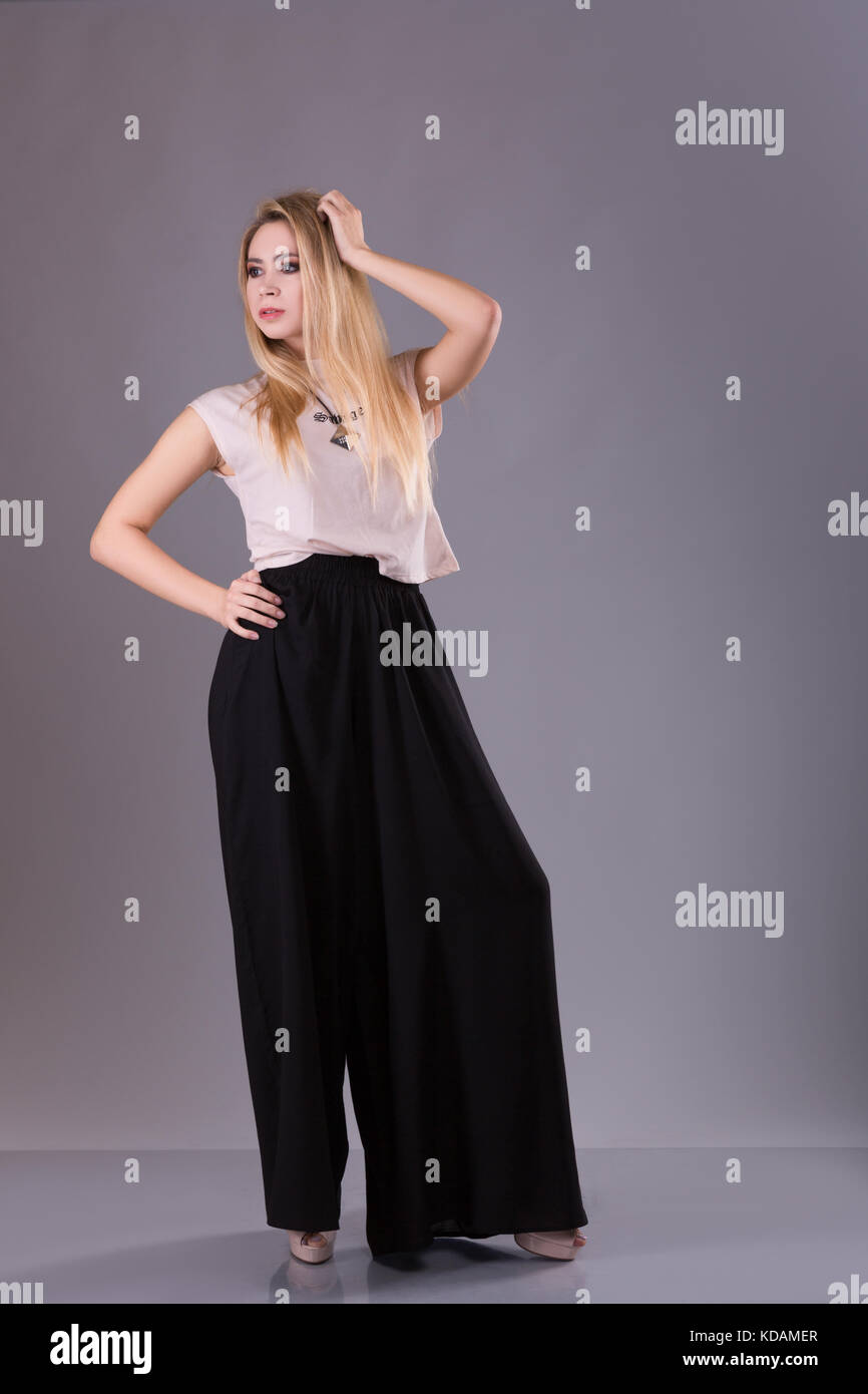 Young blond woman in white shirt, black pants, and high heels standing with hand on head. Full length studio shot on gray background. Stock Photo