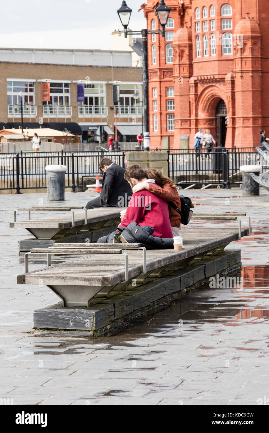 A couple sit on a bench and cuddle, looking out over Cardiff Bay, Cardiff Dock, in the early spring rainy day, Cardiff, Wales UK Stock Photo