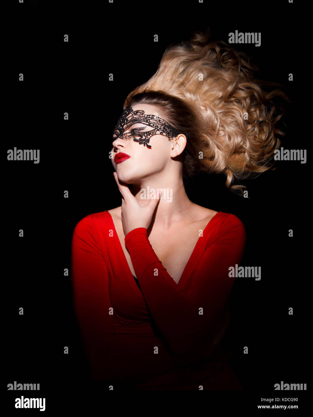 Gorgeous young lady in a red dress, a black mask and with blowing hair over black background Stock Photo