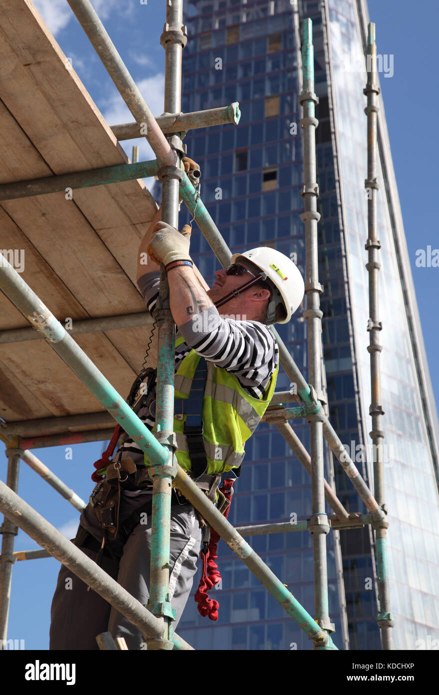 A scaffolder erects a temporary work platform on a tower block high above London. Shows safety harness in use. The Shard is in the background. Stock Photo