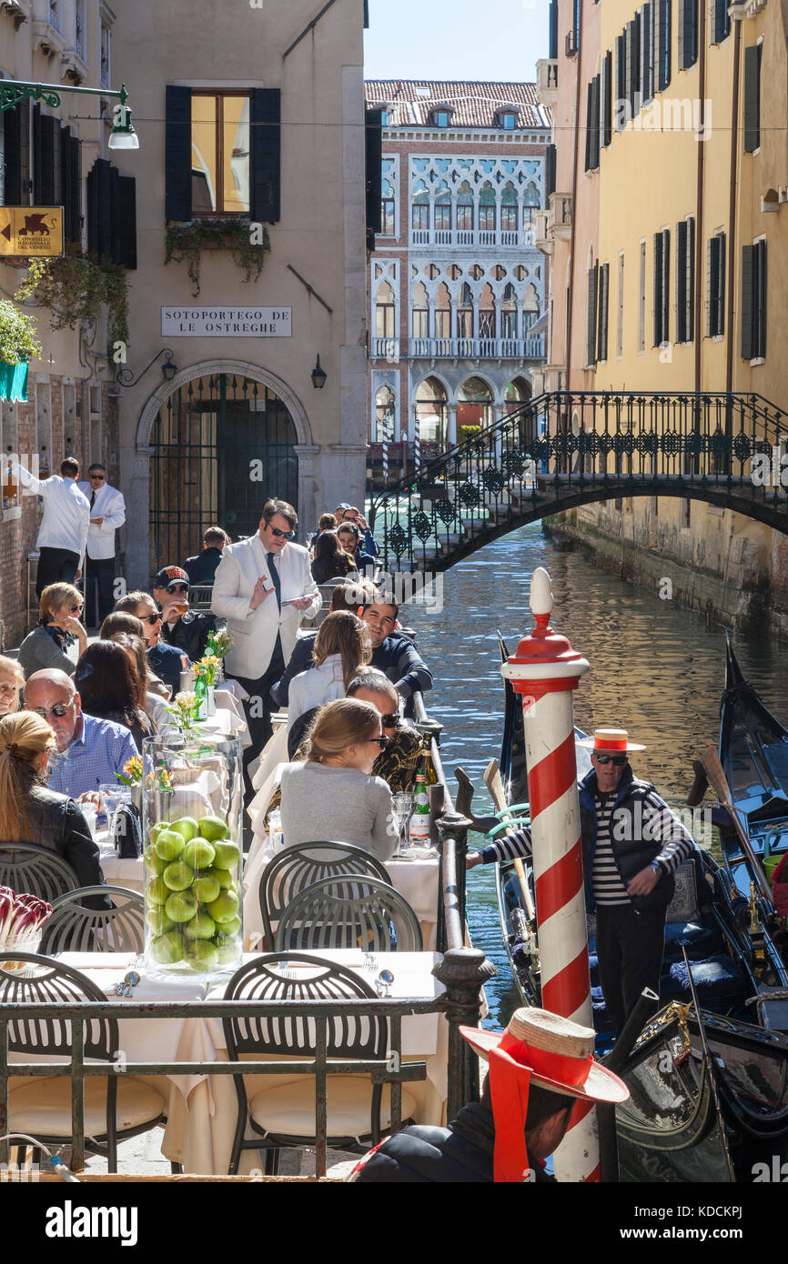 Tourists enjoying the spring sunshine eating  canalside at Ristorante da Raffaele, San Marco, Venice, Italy with gondolas and gondoliers Stock Photo