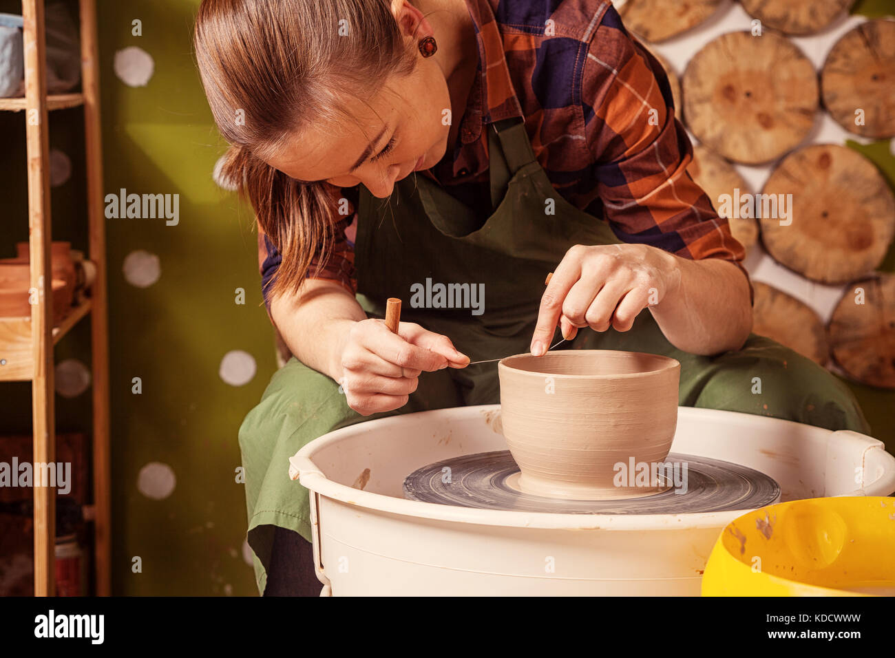 The Dark-haired Woman potter in a plaid shirt and apron sculpts a deep bowl from the clay on a potter's wheel and cuts off excess clay in the workshop Stock Photo