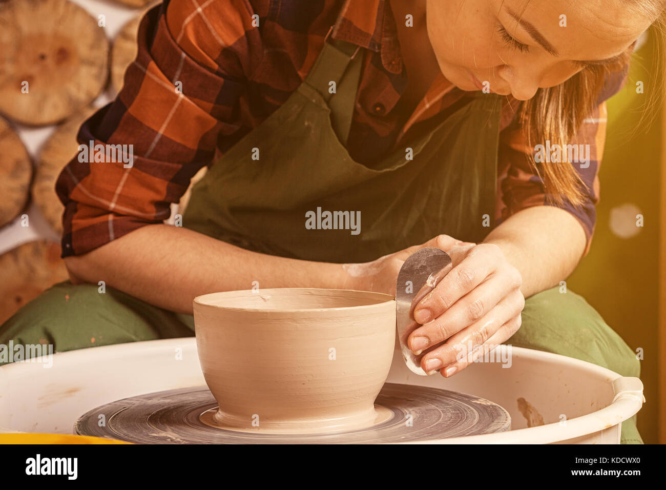 Close-up A dark-haired Woman potter in a plaid shirt and apron sculpts a deep bowl from the clay on a potter's wheel and cuts off excess clay in the w Stock Photo