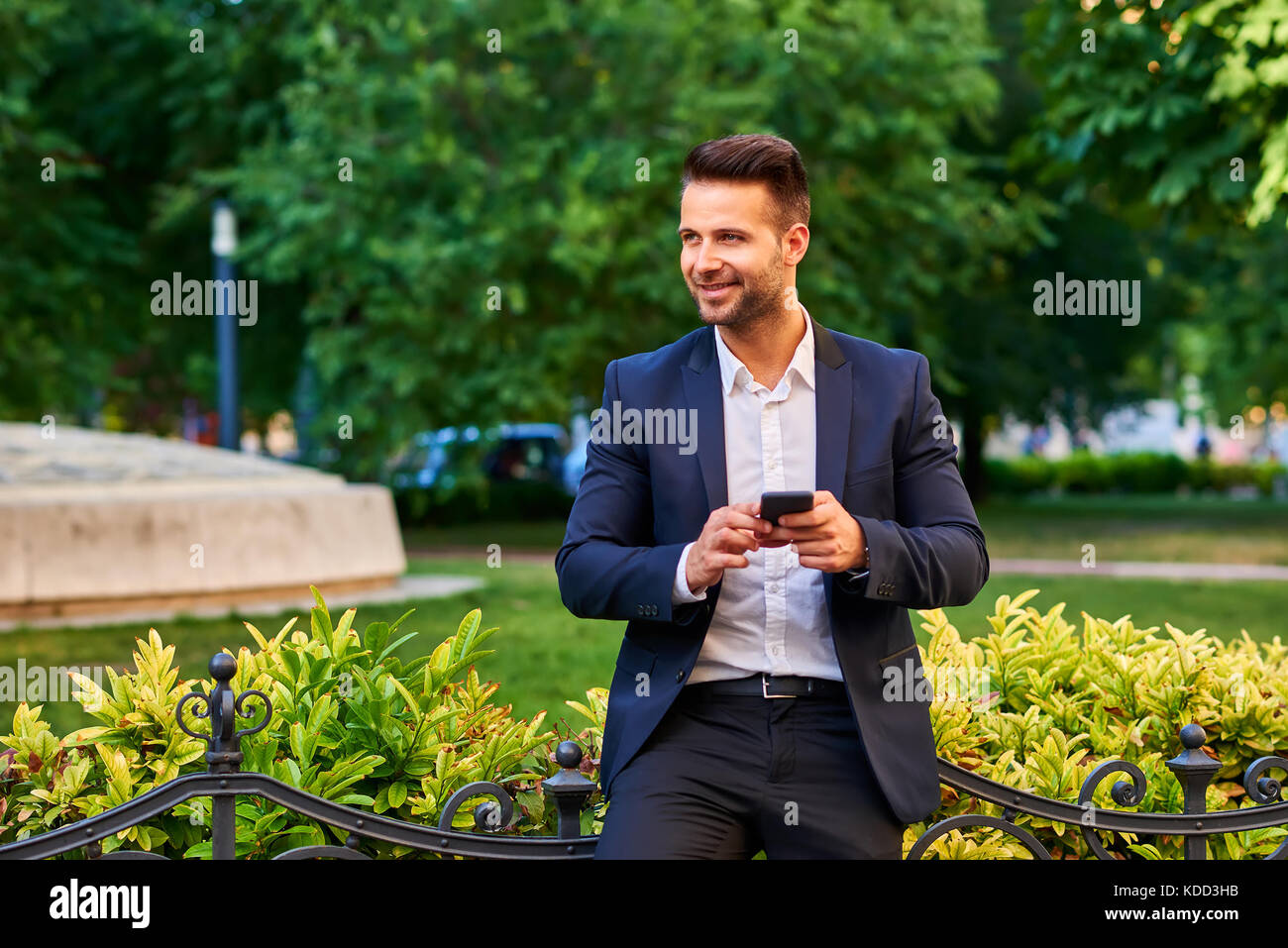 A handsome young businessman leaning against the fence in a park while holding his smartphone Stock Photo