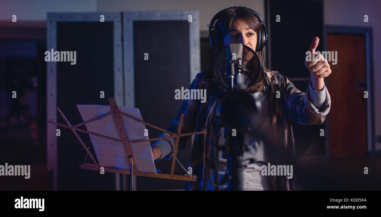 Female singer with microphone and lyrics gesturing thumbs up sign. Woman recording a song for her new album in recording studio. Stock Photo