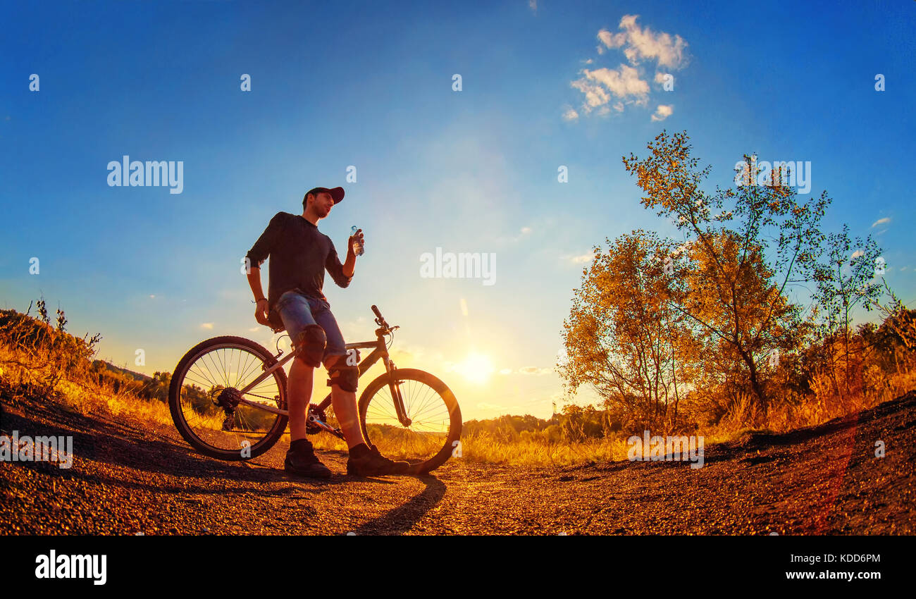 Young athletic guy in a black T-shirt, blue jeans shorts and knee pads on a sports bike holds in hand a plastic bottle with water on an colorful autum Stock Photo