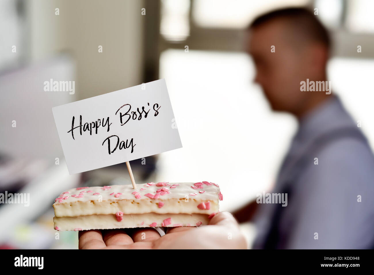 closeup of a piece of cake with a signboard with the text happy boss day written in it placed on a desk, where there is a man working in a computer Stock Photo