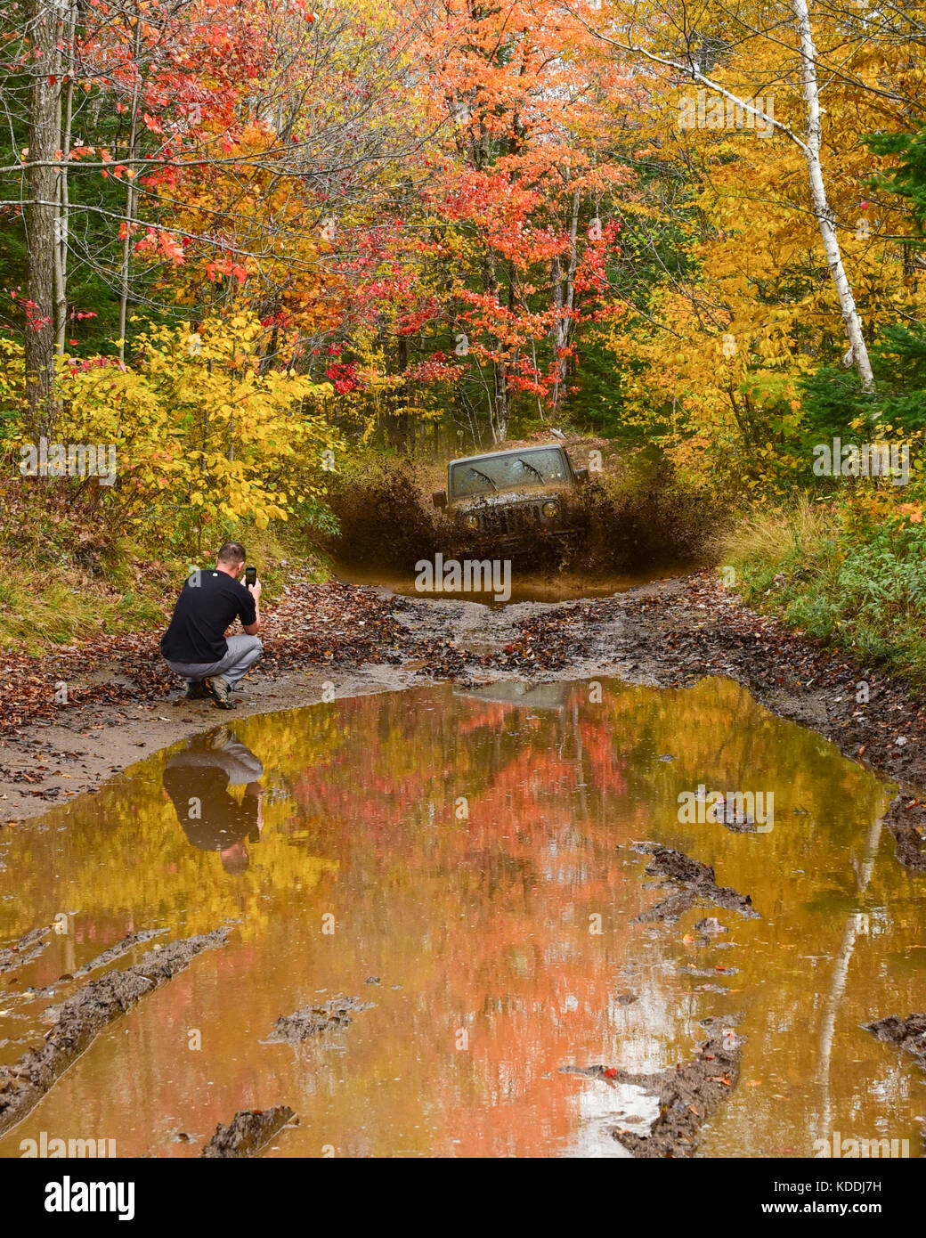 A Jeep Wrangler Rubicon charging through a water and mud hole in the Adirondack Wilderness in autumn, with a photographer taking cell phone pictures. Stock Photo