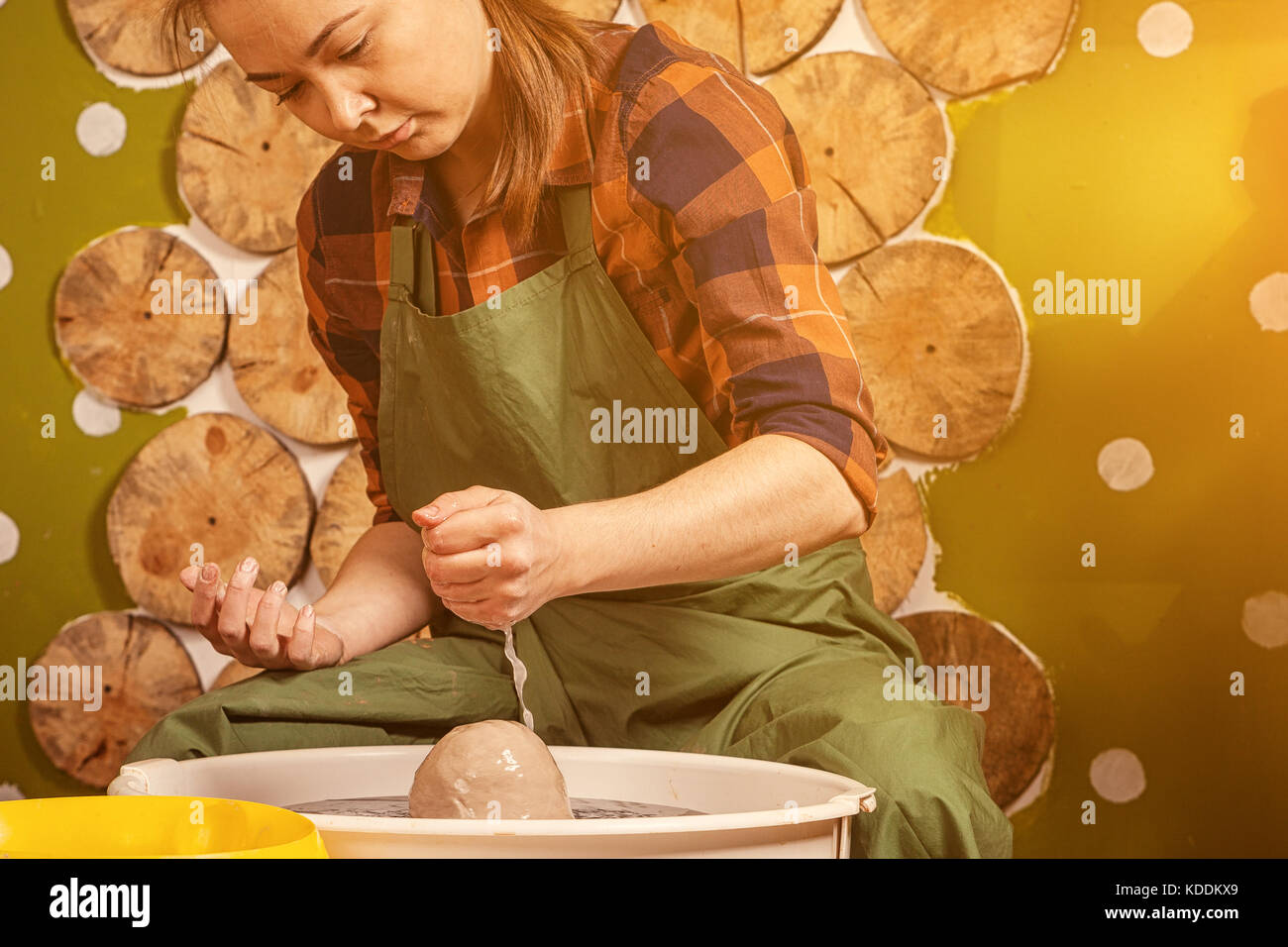 A young woman in a plaid shirt and jeans pours water on the clay to soften it and make a bowl on a potter's wheel in a bright, beautiful workshop deco Stock Photo
