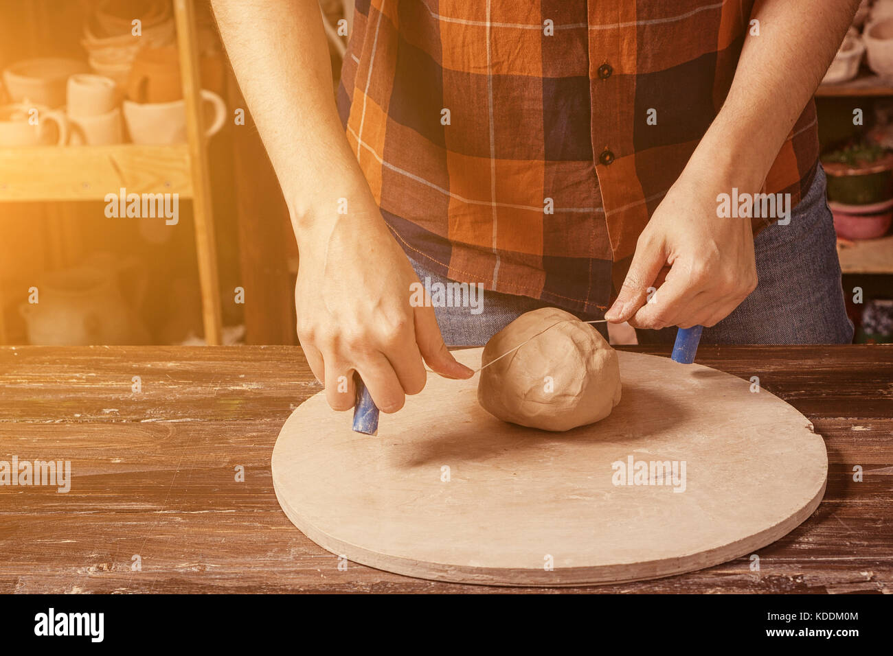 A young woman in a plaid shirt and jeans cuts a glossy ball on a smooth part on a wooden circle in the workshop, in the background a rack with clay po Stock Photo