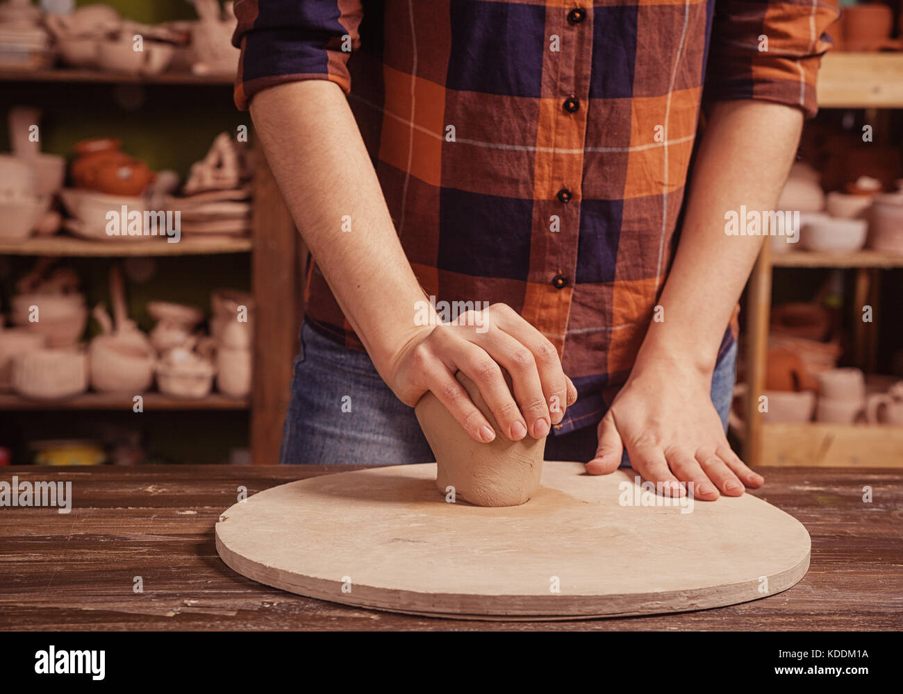 A young woman in a plaid shirt and jeans kneads the clay   and makes a smooth ball before working on a pottery circle of a plate on a wooden circle in Stock Photo