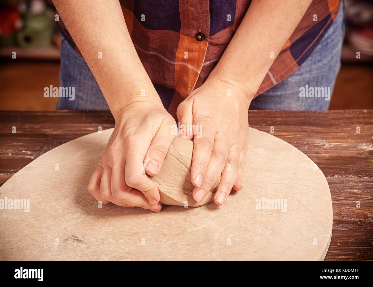 A young woman in a plaid shirt and jeans kneads the clay with her hands before working on the pottery circle of a plate on a wooden circle in the work Stock Photo