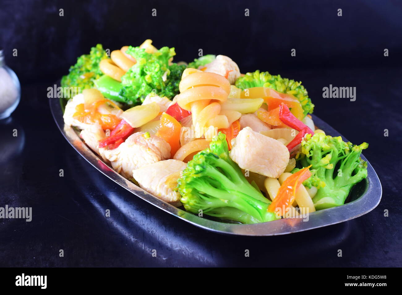 Stir fried chicken fillet with vegetables and pasta on a metal tray on a black abstract background. Stock Photo