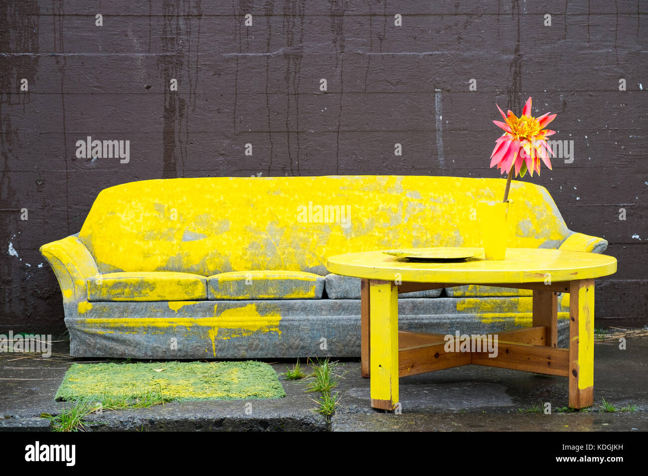 old sofa covered with yellow paint with a table and fake flower in front of it set against a brown wall Stock Photo