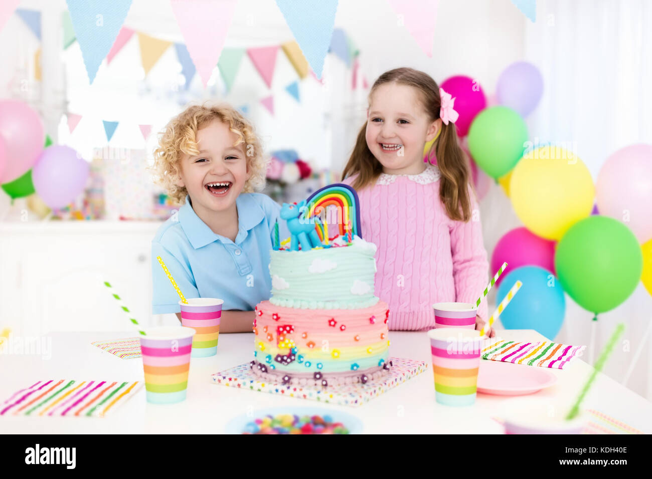Kids birthday party with colorful pastel decoration and rainbow cake. Girl and boy with sweets, candy and fruit. Balloons and banner at festive decora Stock Photo