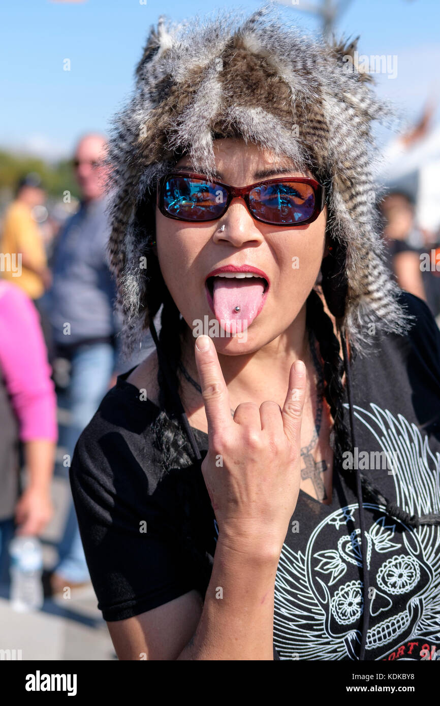 Port Dover, Ontario, Canada, 13th October 2017. Thousands of motorcyclists from all over Canada and the United States get together for The Friday 13th Motorcycle Rally, held every Friday the 13th in Port Dover, Ontario, Canada, since 1981. The event is one of the largest single-day motorcycle events in the world. The mild weather contributed for a large crowd, with hundreds of custom motorcycles, vendors, live music and interesting people to watch. An Asian mid-thirties woman wearing a feather hat makes a hang ten sign while showing her pierced tongue. Credit: Rubens Alarcon/Alamy Live News Stock Photo