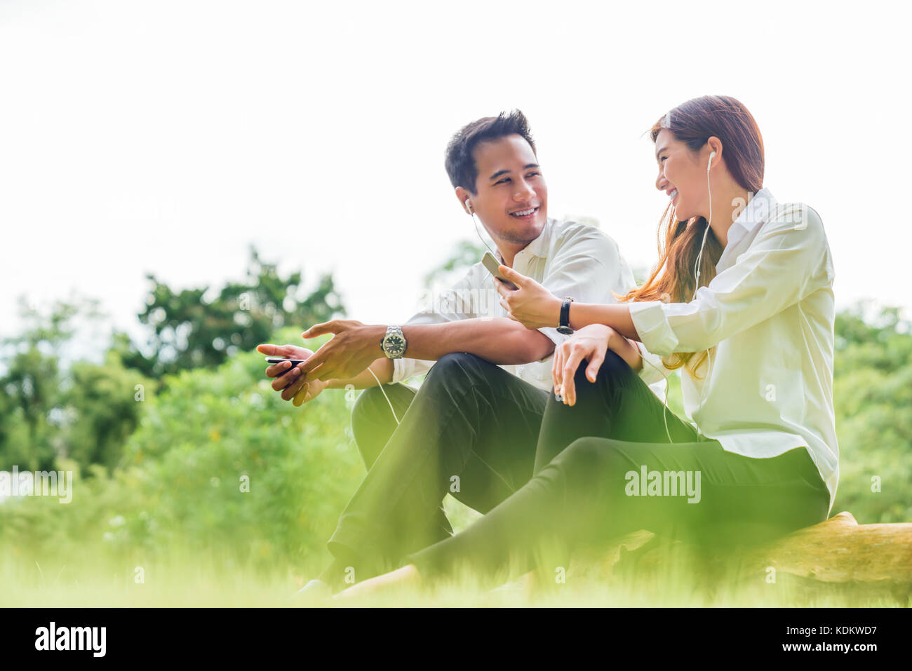 Young Asian lovely couple or college student sit listening to song music on smartphone together in park. Leisure activity, dating relationship concept Stock Photo