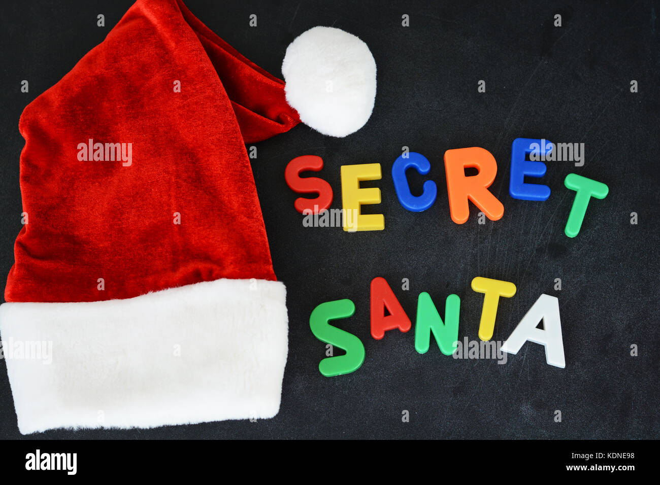 Secret Santa text with colorful magnetic letters on blackboard near Santa’s red hat Stock Photo