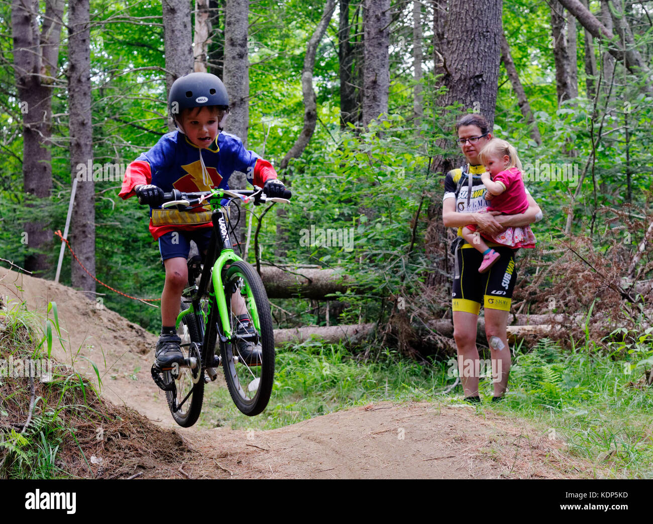A young boy (5 yrs old) jumping in a mountain bike, while mum holding the sister, watches on Stock Photo