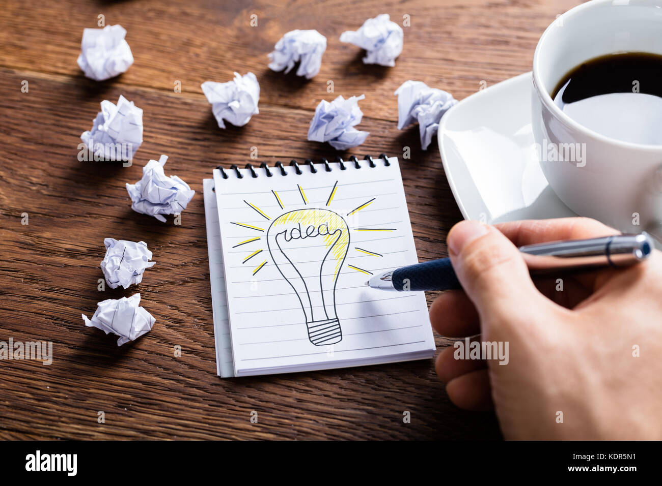 High Angle View Of Person Drawing Lighting Bulb On Notepad Near Crumpled Paper On Wooden Desk Stock Photo