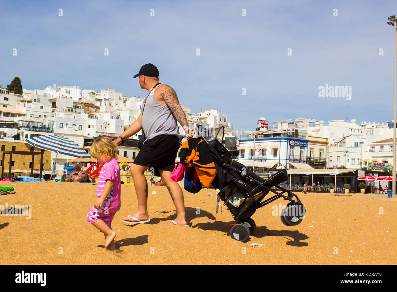 A young father with his child pulling a push chair across the deep sand on the beach at the Old Town in Albuferia Portugal Stock Photo