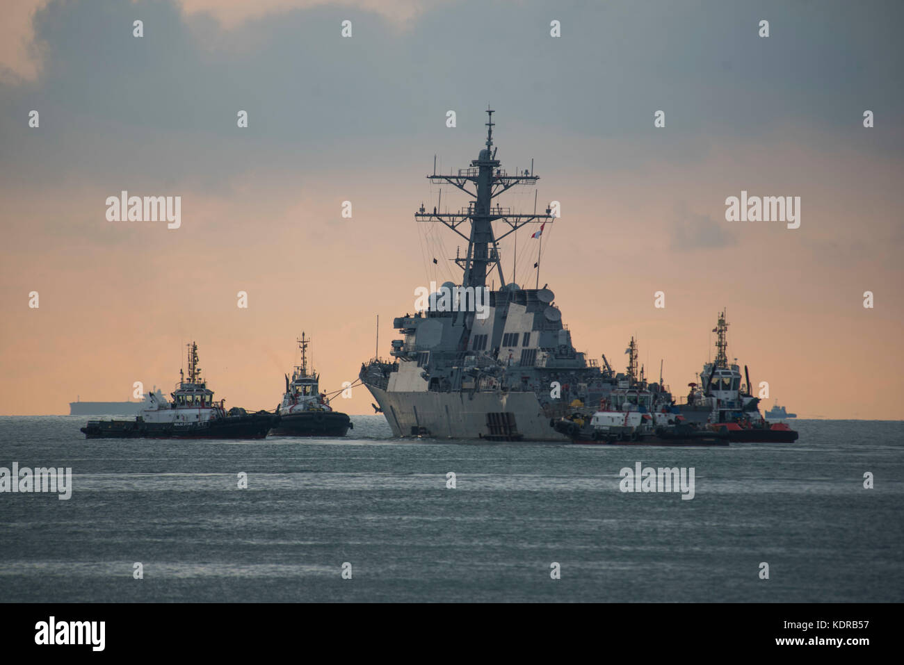 A tug boat tows the U.S. Navy Arleigh Burke-class guided-missile destroyer USS John S. McCain away from the Changi Naval Base pier at sunset October 5, 2017 in Changi, Singapore. Stock Photo