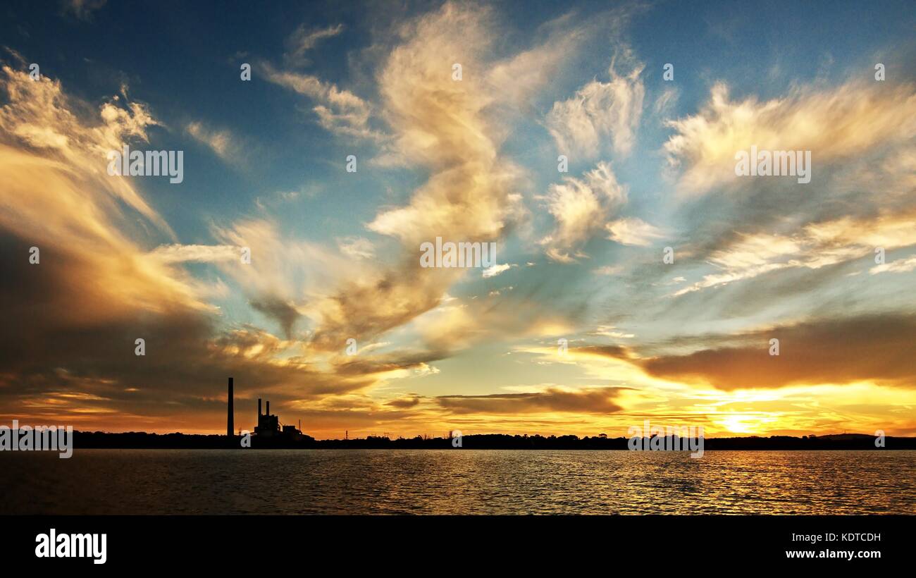 A striking inspirational gold coloured cloudy salt water lake sunset over a power station with water reflections. Captured on Lake Macquarie, Australi Stock Photo