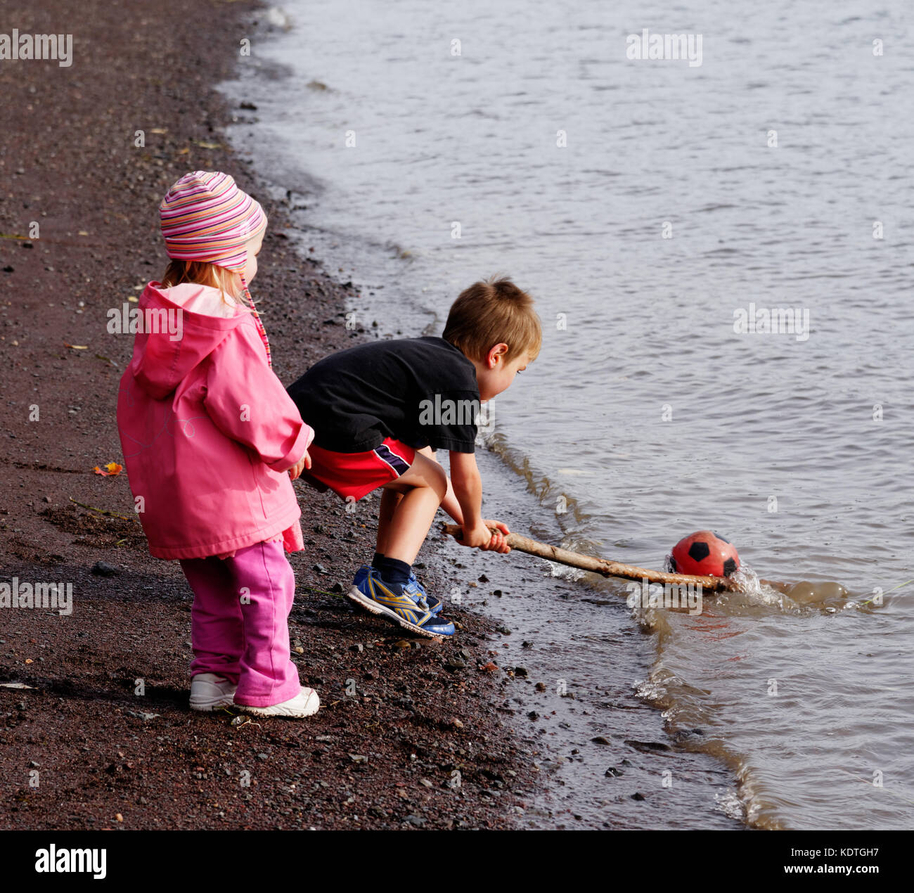 A little boy using a stick to rescue his sister's ball that has gone into the sea Stock Photo