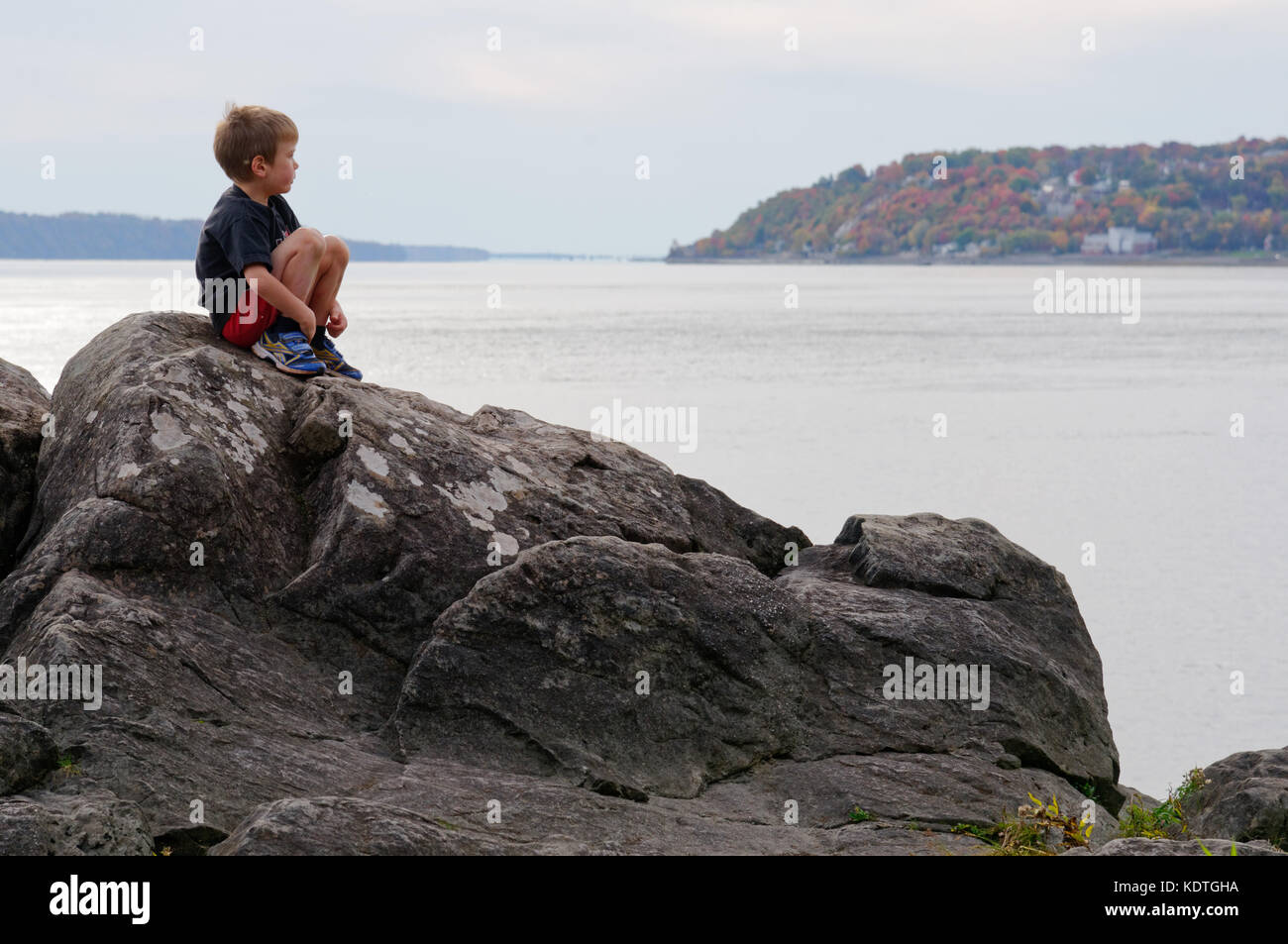 A little boy (5 yrs old) sitting alone on rocks by the River St Lawrence at Cap Rouge near Quebec City, looking thoughtful Stock Photo
