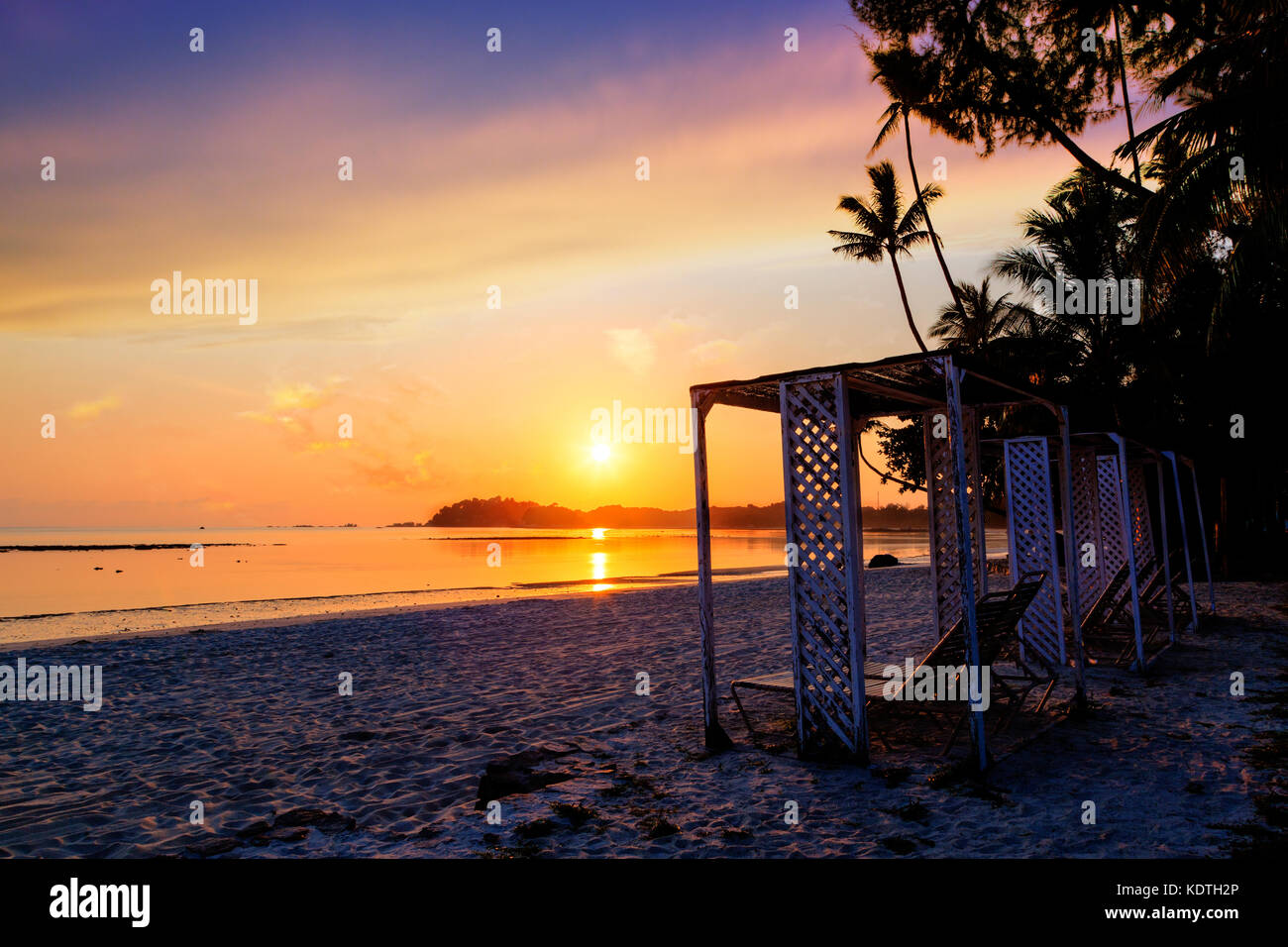 Beautiful golden sunrise over sandy beach on Bintan Island in Indonesia with silhouette of lounge chairs and coconut palm trees with copy space. Stock Photo