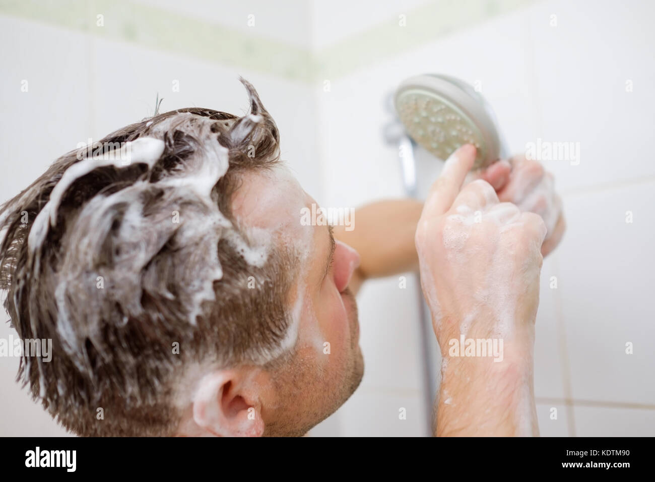 A young man with a soapy head is standing in the bathroom and looks at the shower with perplexity. Stock Photo