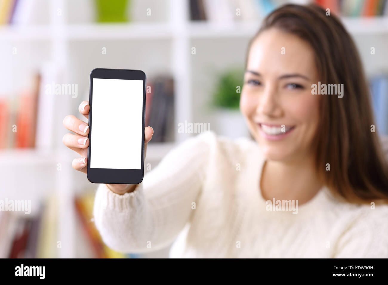 Woman wearing sweater showing a smart phone screen sitting on a sofa at home Stock Photo