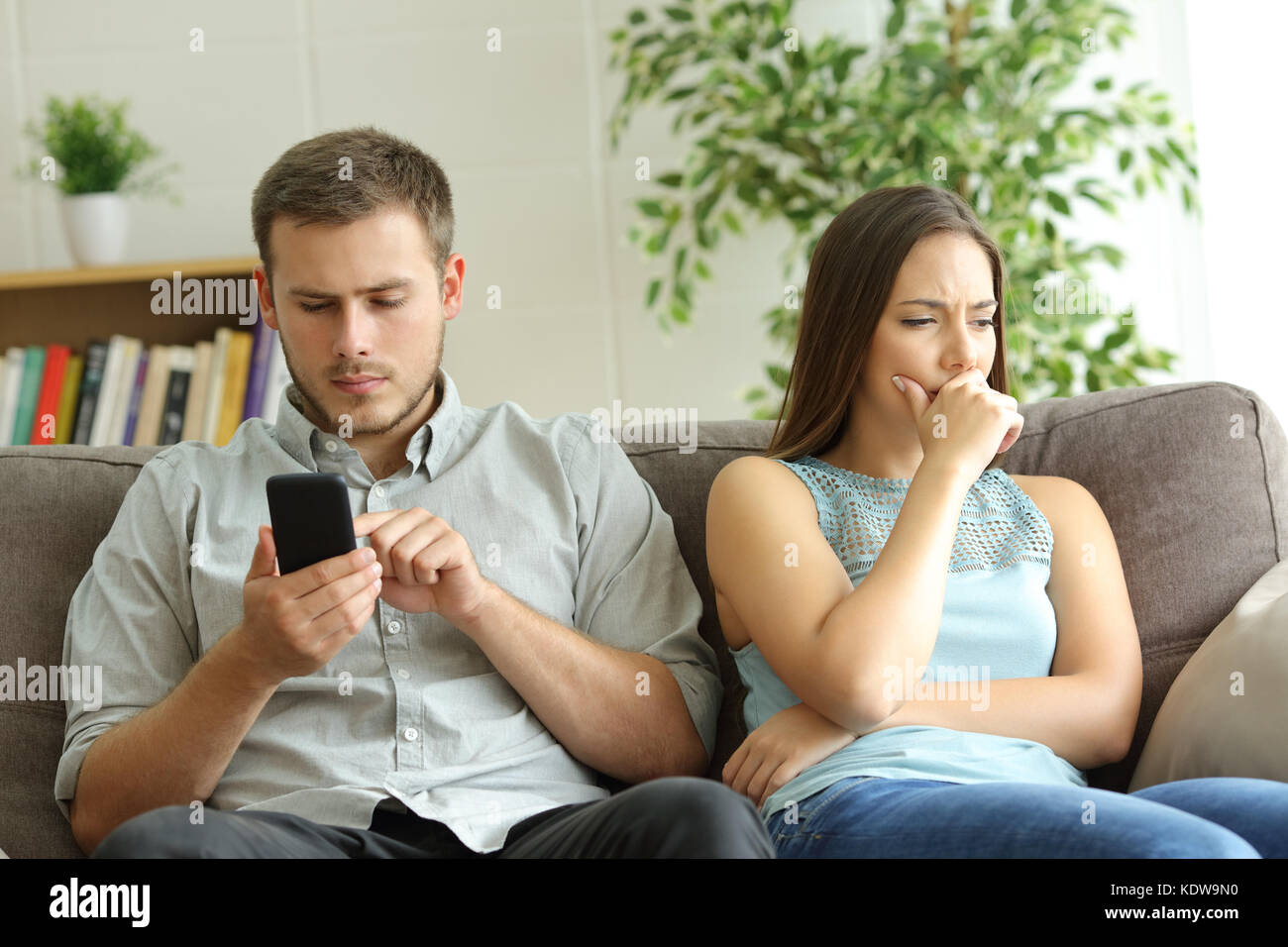 Husband addicted to smart phone watching content beside his worried wife looking down sitting on a sofa at home Stock Photo