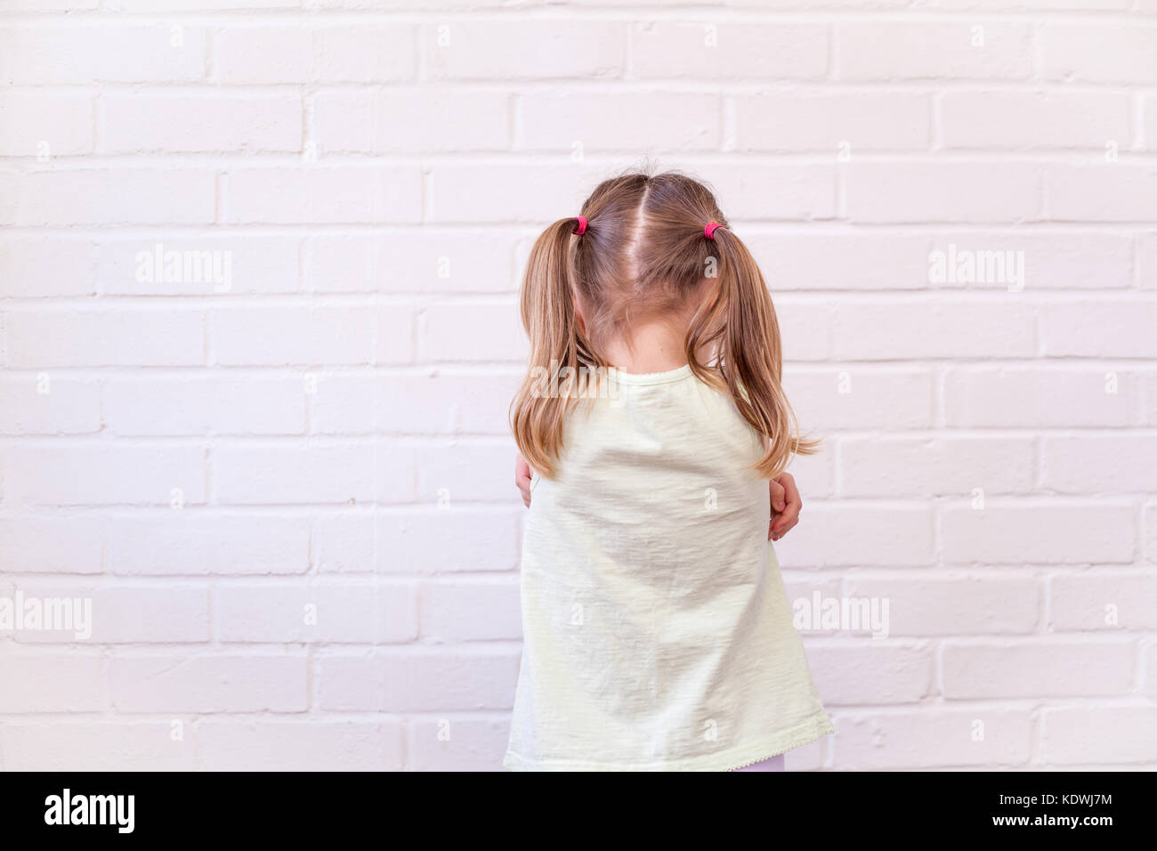 Female child with back to viewer and arms folded in grumpy or sad pose against white brick wall. Emotional or unhappy kid concept Stock Photo
