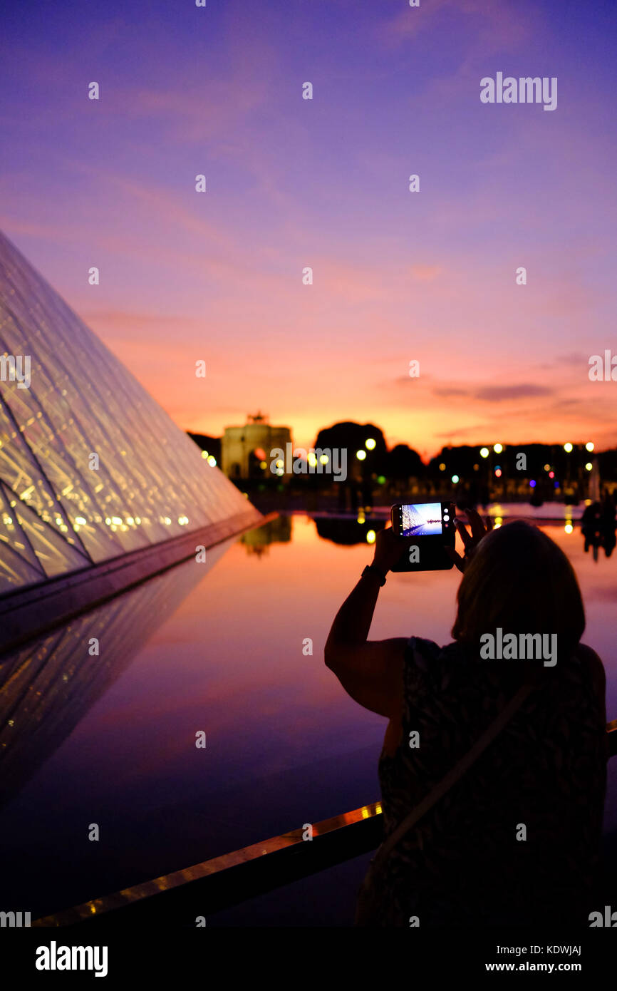 A woman tourist takes a photo of the Louvre at night in Paris, using her smartphone Stock Photo