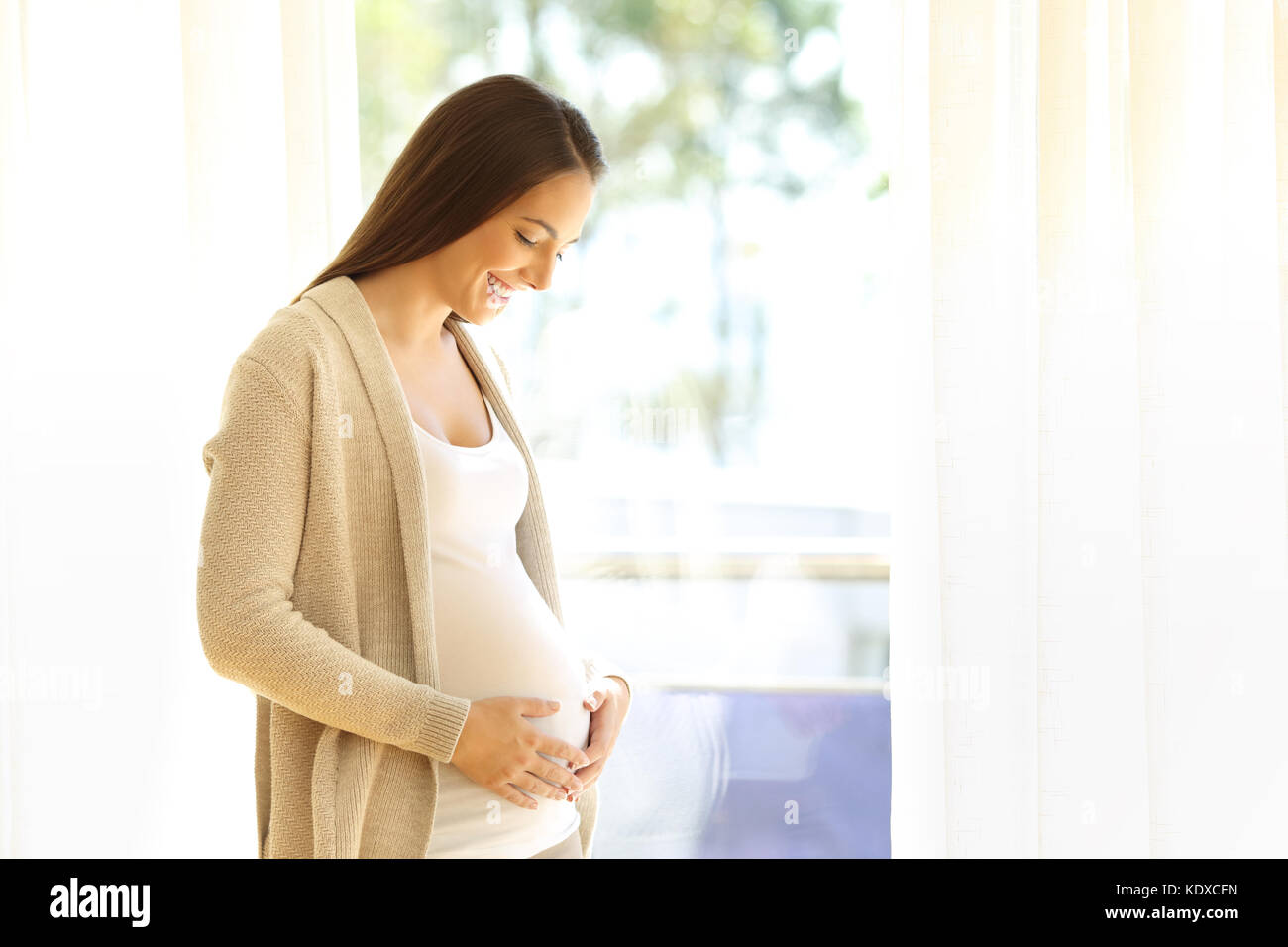 Portrait of a pregnant mother looking at her belly beside a window at home Stock Photo